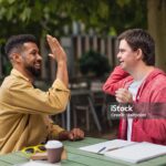 Young man with Down syndrome with his mentoring friend sitting outdoors in cafe high fiving.